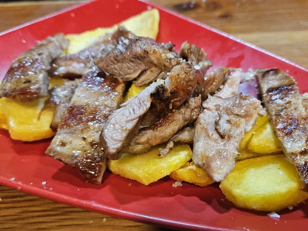 Ración de carne asada cortada en tiras sobre patatas, con sal gruesa, servida en plato rojo en La Taberna del Loco (Algeciras)